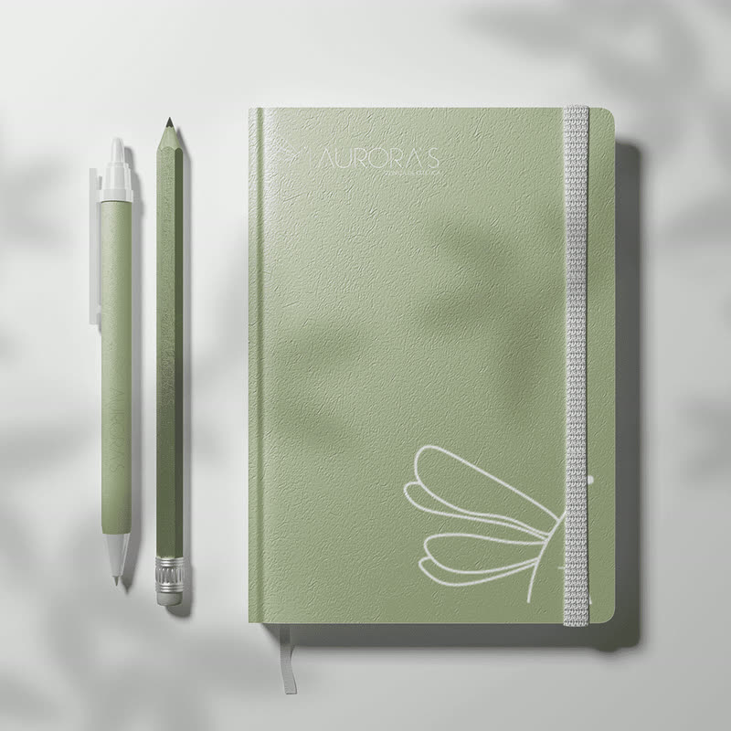 Caderno verde e canetas com a marca da Aurora’s, clínica de estética, e o desenho de libélula